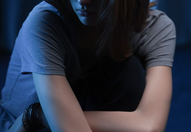 Молодая женщина в серой футболке сидит в полумраке, обхватив колени руками